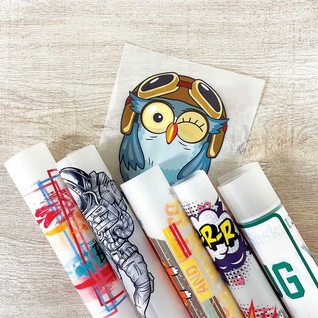 Custom DTF (Direct to Film) heat transfer sheets featuring colorful designs including an owl, astronaut, and comic-style graphics, rolled and ready for application