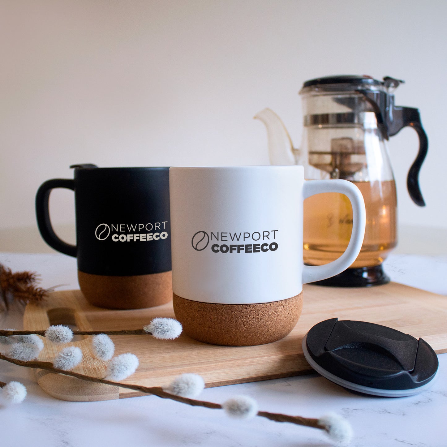 Black and white Newport Coffee Co eco-friendly ceramic mugs with cork base and lids, displayed on a wooden board with a tea pot in the background, ideal for sustainable drinkware promotions
