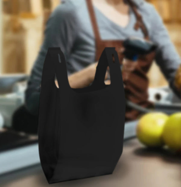 Black T-Shirt style non-woven shopping bag, 10 inches wide by 18 inches high by 5 inches deep, 30 gsm blank, displayed at grocery checkout with fruits and cashier in background.