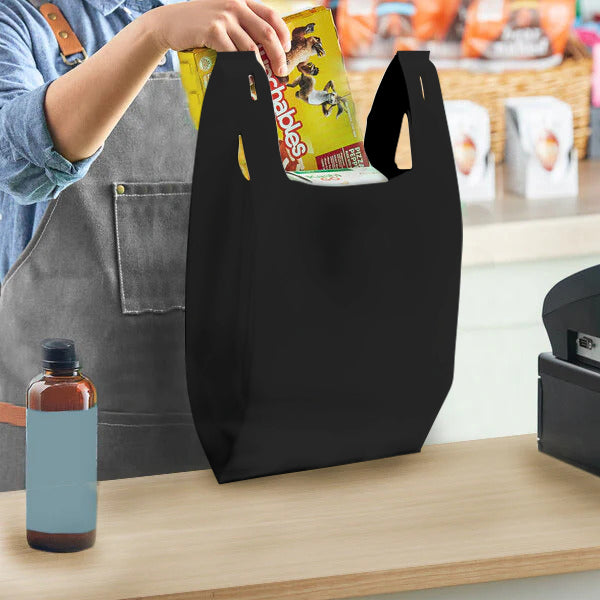 Black T-Shirt style non-woven shopping bag, size 10 inches wide by 18 inches high by 5 inches deep, 30 gsm blank, shown in use at checkout counter holding groceries.