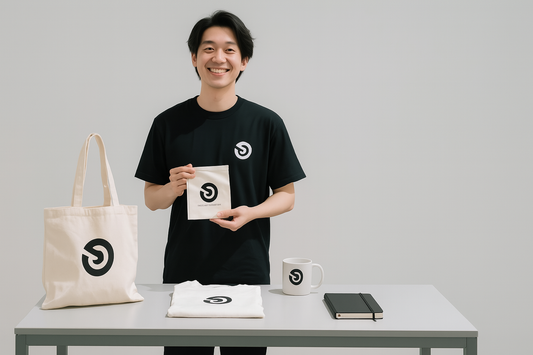 Young small business owner standing in a minimalist studio, smiling while displaying custom-branded merchandise including a tote bag, T-shirt, mug, notebook, and pouch on a clean table.