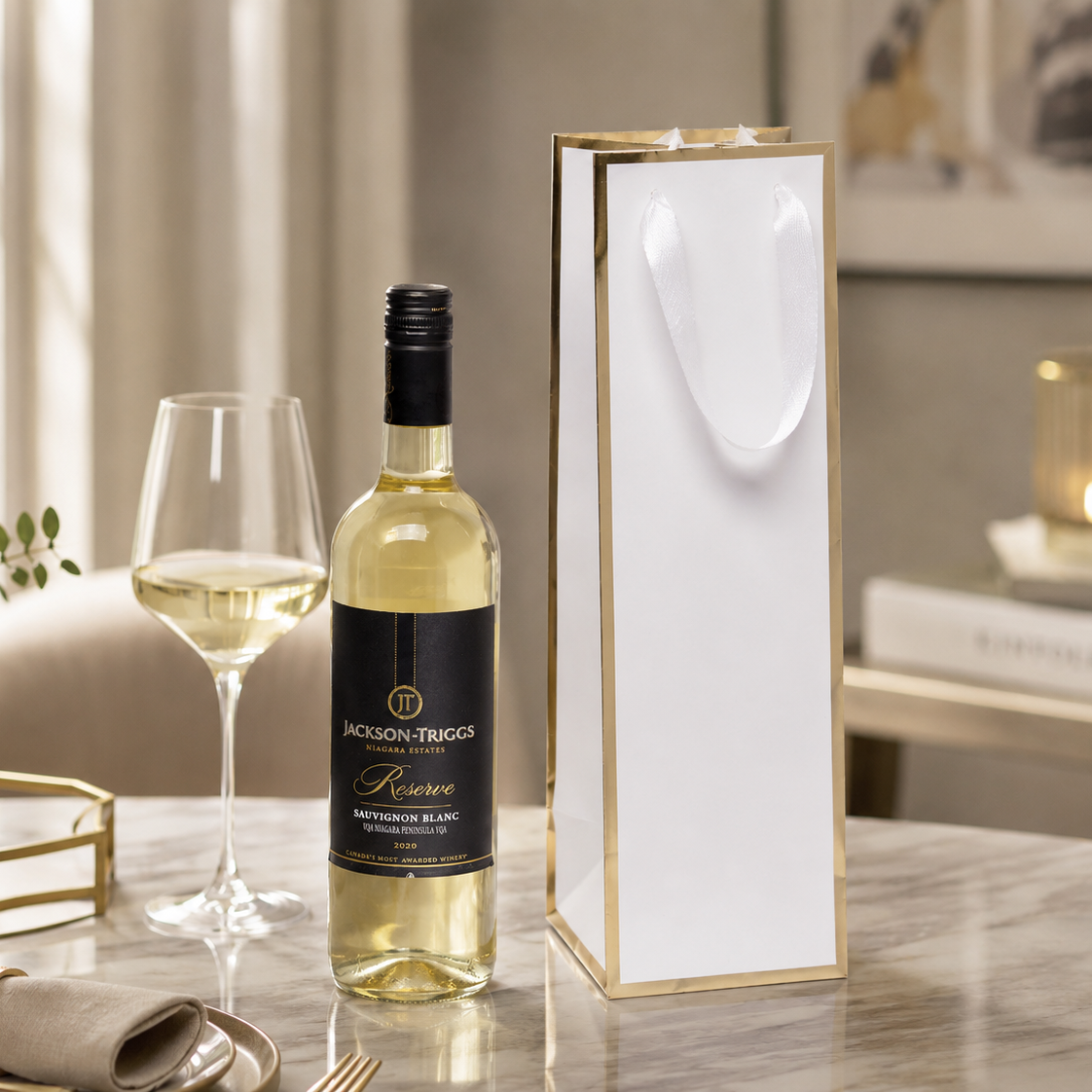 Premium white wine gift bag with metallic gold trim and soft white ribbon handles, displayed beside a bottle of white wine and wine glass in an upscale lifestyle setting. Ideal for wineries, liquor stores, corporate gifting, holiday packaging, and promotional wine packaging. Manufactured by a professional custom wine bag factory offering wholesale paper wine bags with custom printing and branding options.