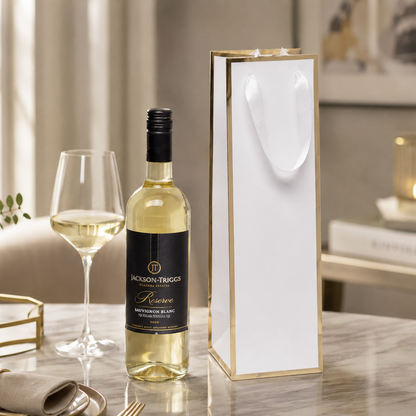 Premium white wine gift bag with metallic gold trim and soft white ribbon handles, displayed beside a bottle of white wine and wine glass in an upscale lifestyle setting. Ideal for wineries, liquor stores, corporate gifting, holiday packaging, and promotional wine packaging. Manufactured by a professional custom wine bag factory offering wholesale paper wine bags with custom printing and branding options.