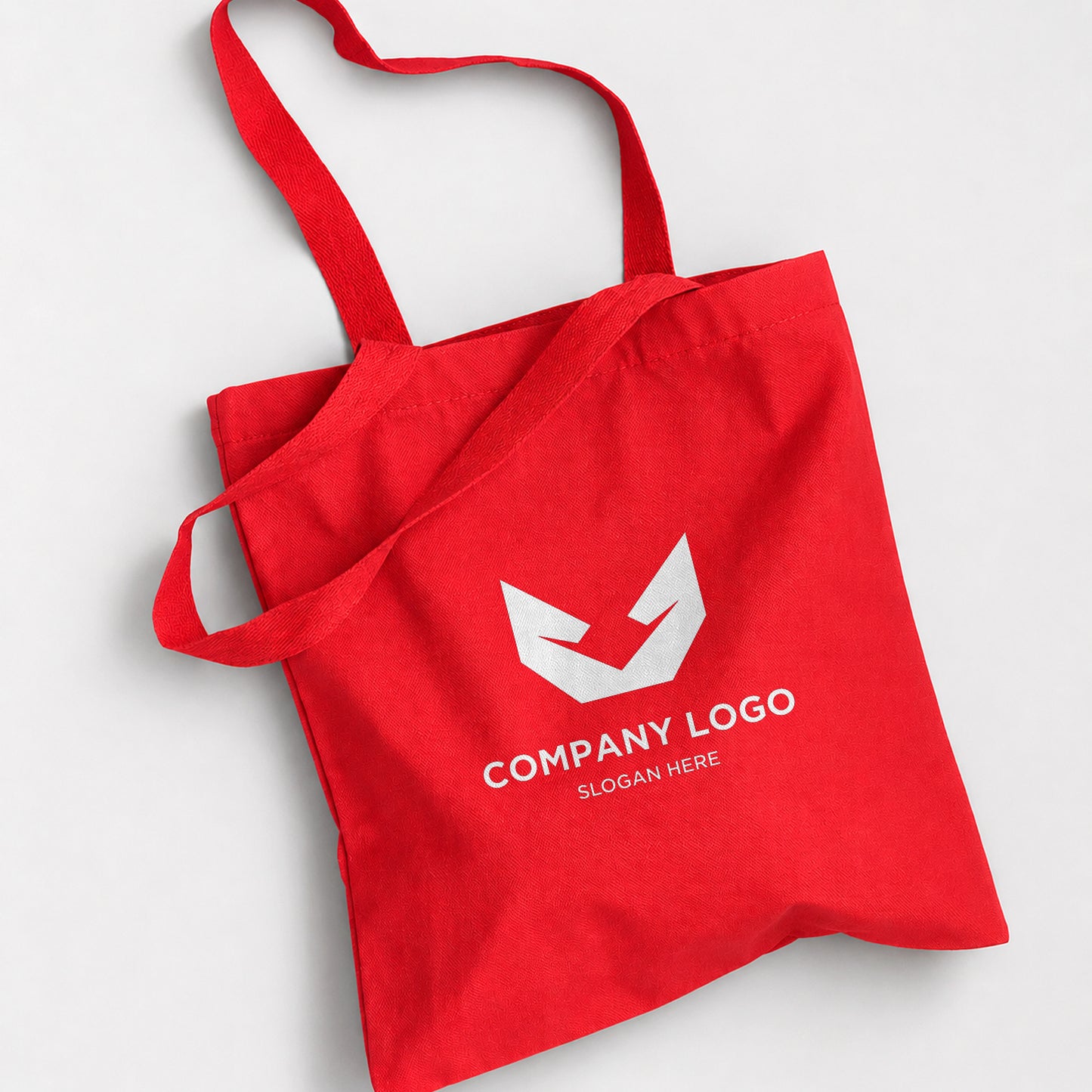 A red canvas tote bag with two shoulder straps, featuring a white geometric logo and the placeholder text 'COMPANY LOGO' and 'SLOGAN HERE' printed on the front, set against a light gray background.